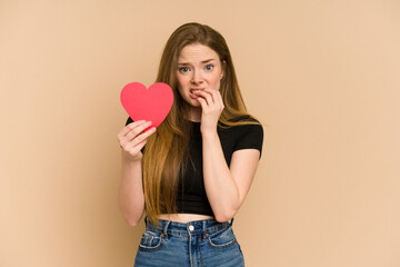 Young redhead woman holding a paper red heart cut out isolated biting fingernails, nervous and very anxious.