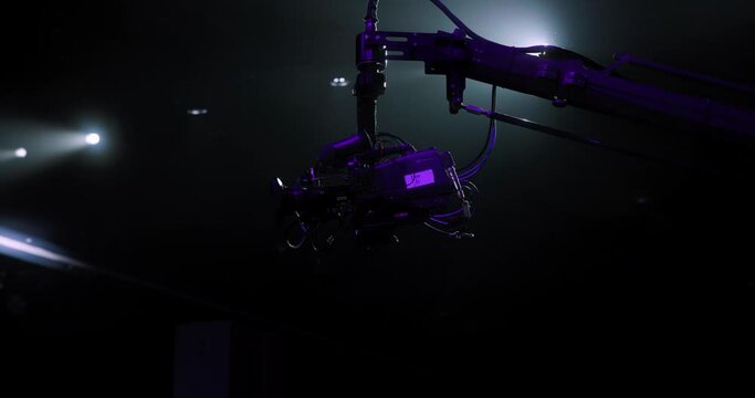Professional production camera on long crane arm operating under dramatic spotlight and moving.