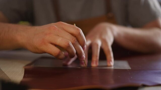Leathersmith draws outline on leather sheet with plastic mould at workplace in home studio closeup. Master makes item of natural material. Art hobby