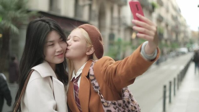 Lesbian couple kissing and gesturing while taking a selfie outdoors