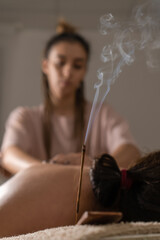 Close-up of smoking incense, in the background, a masseur doing a back massage in a cozy office. Concept of health, body care, Vertical photo