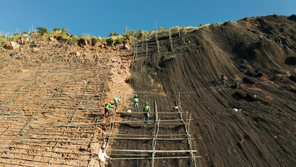 Protection of road from mountain slough, rockfall with metal accumulative restraining net fences. Workers constructing anti-landslide concrete wall prevent protect against rock slides. Rockfall