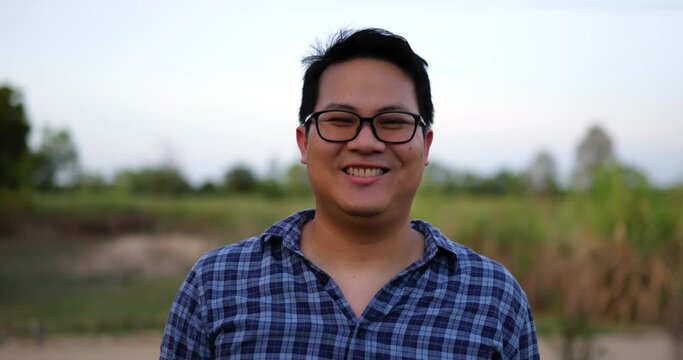 Portrait of an Asian young happy farmer man with glasses looking at camera and smiling with a farmland background. Agriculture farming concept. Slow motion.
