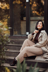a brunette woman in a raincoat sits on the steps on the streets of autumn city.