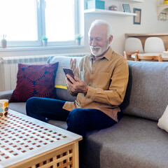 Grey-haired senior hipster man in casual shirt using smartphone sitting on the sofa at home. Elderly guy enjoys online chatting, spends time in social networks, using phone and smiles.