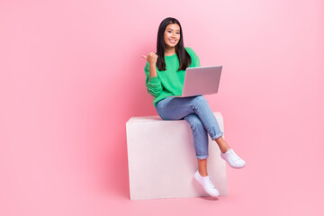 Full length photo of charming cheerful person sit podium use netbook indicate finger empty space isolated on pink color background