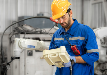 Male industrial engineer using remote control board to check robotic welder operation in modern automation factory. Factory technician monitoring robot controller system for automated steel welding.