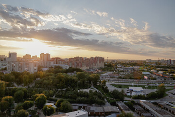 Fototapeta na wymiar Sunset illuminates busy city with residential area surrounded by park with green trees and road. Sunlight breaks through cumulus clouds