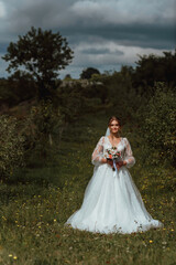 a young beautiful bride poses with a wedding bouquet against the background of nature; the girl is dressed in a white wedding dress; a moment on the wedding day; happy girl expresses her emotions