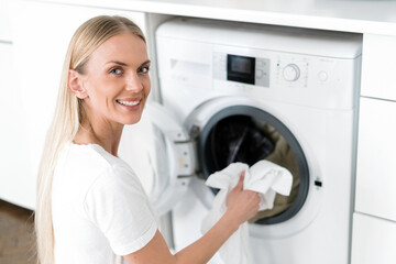 happy female housewife load laundry in washer appliance at home