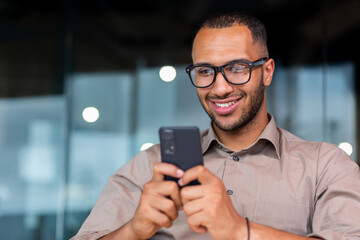 Obraz na płótnie Canvas Close-up of smiling African-American businessman using phone typing messages and browsing internet pages, man in shirt and glasses smiling happily inside office holding smartphone.