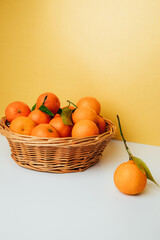 Orange mandarins clementine with green leaves in straw basket on a white-yellow background. Front view