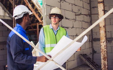 Two diversity male engineers team working, inspecting outdoor at construction site, wearing hard hats for safety, talking, discussing, holding plan for building. Career, Industry Concept.