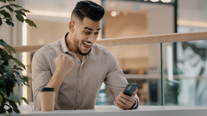 Naklejka premium Indian Hispanic man happy shocked amazed male businessman has offer online message good news excited winner guy win victory financial prize with smartphone lucky celebrating winning with mobile phone