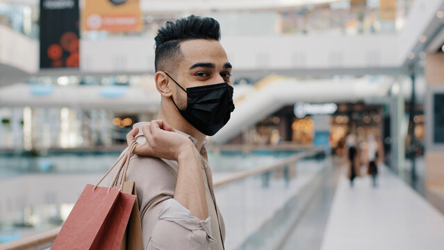 Masked Stylish Ethnic Guy Indian Hispanic Man In Face Medical Mask Businessman Buyer Male Client Customer In Shopping Mall Turning Back Posing With Purchases Holding Bags Gifts Packages Discounts Sale