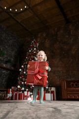 Obraz premium Little girl with big gift on xmas tree background. Cute blonde child girl with huge red gifts box.