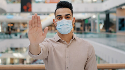Fototapeta na wymiar Portrait of young hispanic man wearing medical mask put palm forward serious ethnic guy looking at camera male showing stop sign palm with hand gesture safe distance coronavirus epidemic protection