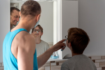 Young father and his teenage son with shaving foam on his nose. Dad and son in bathroom in front of mirror together.