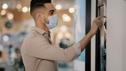 Hispanic Indian passenger male buyer man in medical mask in airport cafe touching self-service machine in buying ticket make online order scroll touch screen panel. Businessman using terminal display