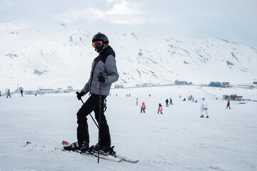 Fototapeta na wymiar A girl in a ski suit, a helmet and a sun mask stands on skis and with ski poles on a mountain slope against the backdrop of snow-covered mountains. Winter. Extreme sport and travel content