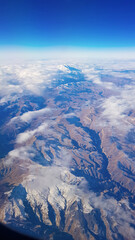 View from window of plane to blue sky and snowy mountain peaks in white clouds. High quality photo