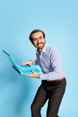 Cheerful Caucasian man, businessman in office dress code having fun, dancing with laptop over blue background. Positive emotions, work, happiness