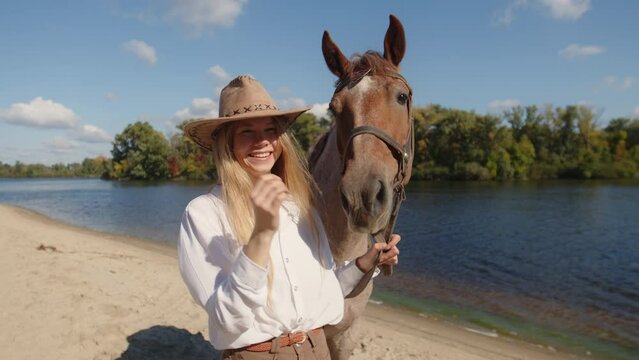 Woman walking beautiful animal towards the clear blue pond. Close-up view of a charming, smiling girl wearing a hat and pets a horse. High quality 4k footage