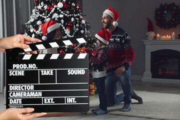 Shooting movie. Second assistant camera holding clapperboard in front of happy family celebrating Christmas (actors) at home (film set) - obrazy, fototapety, plakaty