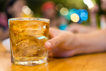 A glass of scotch whiskey with ice held by woman against a colored bokeh background at pub