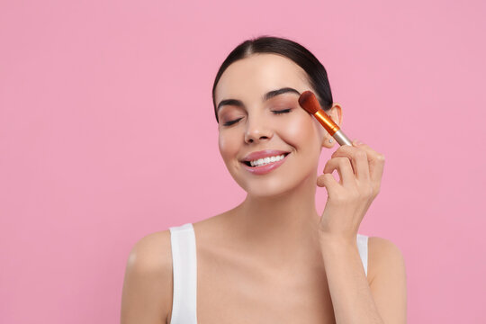 Beautiful woman applying makeup with brush on pink background. Space for text