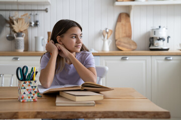 Dreamy optimistic teenage girl sits at kitchen table with books distracted from doing...
