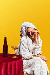 Young woman in bathrobe sitting and drinking champagne over yellow background. Celebration