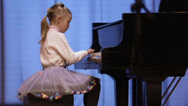 Cute 7 years old girl playing piano on stage in concert under the light of colored soffits. A casual day at a music school