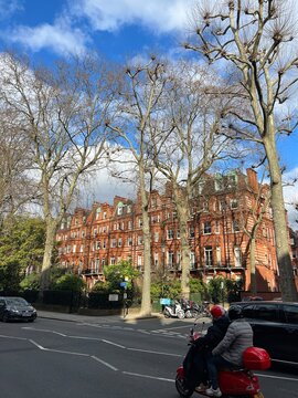 A couple on a red scooter passing by a Chelsea red brick victorian building