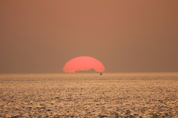a ship sails in front of the last part of the red sun just above the sea water during sunset at the coast in summer