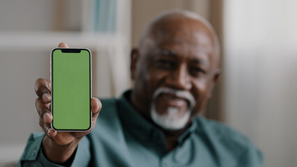 Old male African American biracial man at home sitting on couch businessman in office showing smartphone with empty green screen chromakey holding phone shows to camera advertisement gadget touchpad