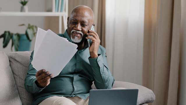 African biracial bald old man with gray beard senior mature businessman busy employee using laptop talking cell phone in multitasking discussing online project distant paper documents remote from home