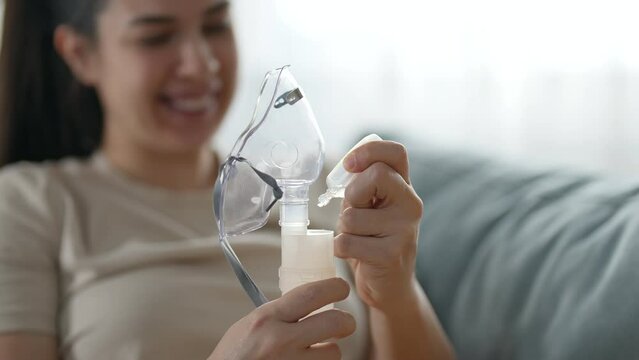 Smiling young woman whose hands are in focus is preparing medicine for inhalation with a mask while sitting on a sofa at home. Pouring the medicine in a nebulizer container.