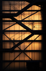 A staircase with several floors is surrounded by a glass house. The sun shines through the glass house and glows orange. The shadows from the metal can be seen, in portrait format