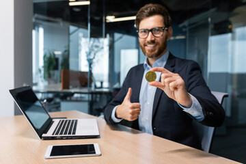 Portrait of mature successful businessman investor, man shows cryptocurrency gold coin to camera and thumbs up firmly, man works on stock exchange inside office online uses laptop.