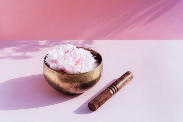 Tibetan singing bowl with floating in water pink peony inside and wooden stick on the pink background. Meditation and Relax. Exotic massage. Minimalism. Direct sunlight and shadows. Selective focus