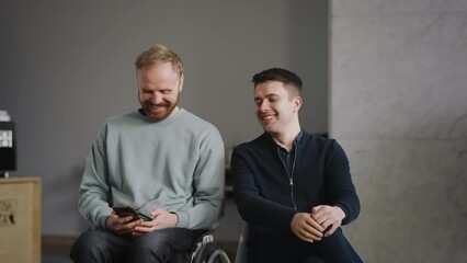 Two disabled men in good mood watching funny video on smartphone indoors. Young caucasian man with silicone prosthesis hand, man on wheelchair. People with physical disability, inclusion concept