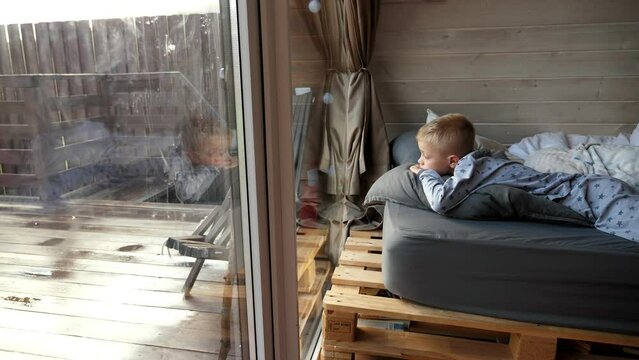 A little boy lies on a bed in a small triangular wooden house and looks out of a large window. Rest in the autumn forest. The house is a bungalow with a roof descending to the ground.