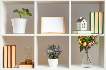 Home interior, elements of Easter decor. White shelves with books, hourglass and plants