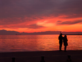 Silhouette shadow of an unidentifiable couple before a vivid color orange sunset by the waterfront and a mountainous range.