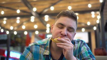 Hungry man is actively eating a burger. A young man in a cafe eats a burger and drinks coffee. Close-up of a man's face.