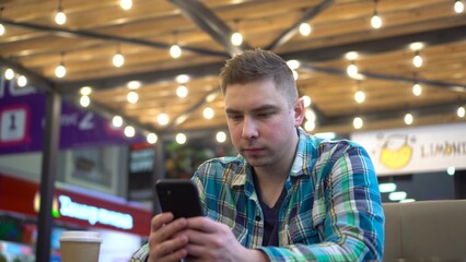 A young man sits in a cafe with a phone in his hands and drinks coffee. A man sits on a food court against the background of cozy light bulbs.