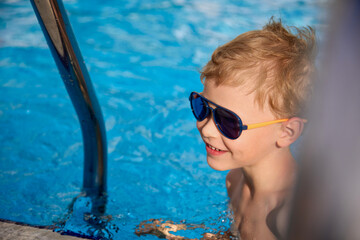 Smiling boy in sunglasses swam up to the steps in the pool to climb. Happy child swims in the warm...