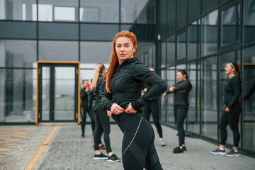 Naklejka premium Redhead is standing against her friends. Group of sportive women is outdoors near black building