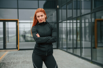 Obraz premium Standing with arms crossed. Woman is sportive clothes is outdoors against black building